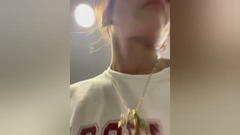 Media: Video of a young woman with light skin, brown hair tied back, wearing a white t-shirt with red text, and a gold necklace with a pendant. Background is blurred with soft lighting.