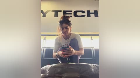 Media: Video of a woman with light skin and brown hair in a bun, wearing a white t-shirt and jeans, sitting on a bus, holding a smartphone.