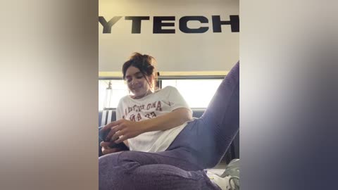 Media: Video of a young woman with dark hair in a ponytail, wearing a white t-shirt and blue pants, sitting on a bed, playing a video game.