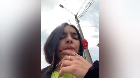 Media: Video of a young woman with long, dark hair, wearing a green top and black jacket, holding a red rose to her mouth, set against a cloudy sky and utility poles.