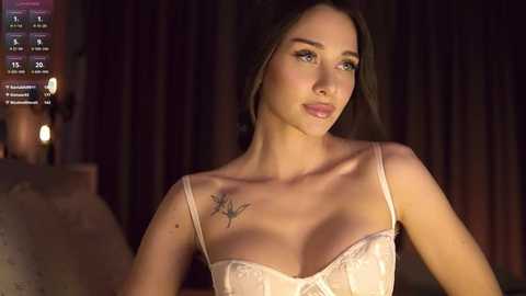 Media: Video of a young Caucasian woman with fair skin and brown hair, wearing a light pink lace bra, with a small star tattoo on her left shoulder. She stands indoors against dark curtains, dimly lit.