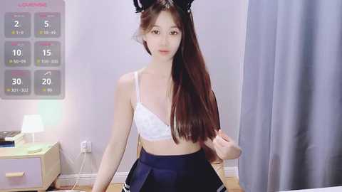 Media: Video of an Asian woman with long brown hair, wearing a white bra and black pleated skirt, standing in a minimalistic bedroom with a calendar and lamp.