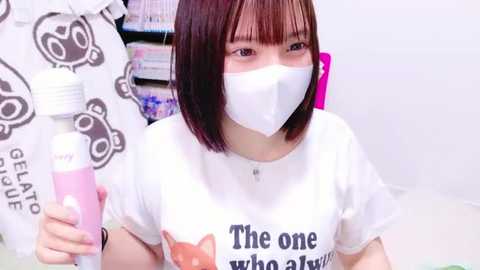 Media: Video of an Asian woman with straight, shoulder-length brown hair, wearing a white T-shirt featuring a fox graphic, and a white surgical mask. She holds a pink hair removal cream and a hair trimmer, in a brightly lit, clean room with bookshelves in the background.