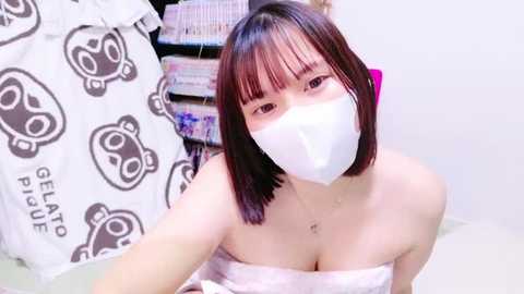 Media: Video of a young Asian woman with straight, shoulder-length black hair and a white face mask, wearing a white strapless top, standing in a cluttered room with a white bed and stacks of books.