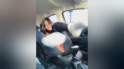 Media: A video captures a woman with dark hair, wearing a blue bodysuit, smiling and playfully kicking her leg, seated in a car. The car's interior is visible with a steering wheel and gearshift.