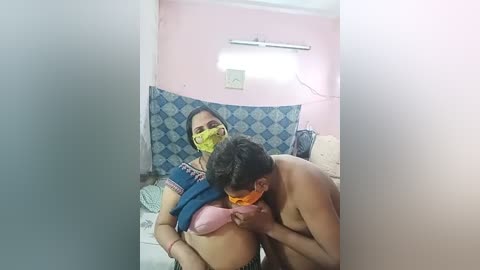 Media: A video captures a topless man kissing a topless woman with a yellow face mask, both sitting on a bed in a pink-walled room with blue patterned sheets and a fluorescent light overhead.