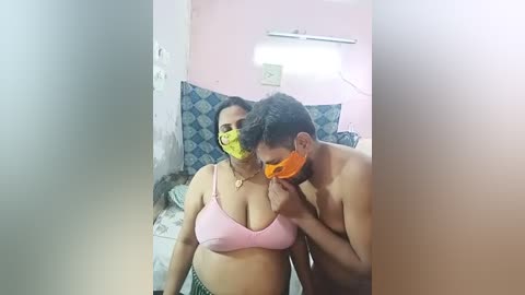 Media: A video captures a topless man with a mask kissing a woman in a pink bra, in a dimly lit room with blue-patterned bed sheets and a pink wall.