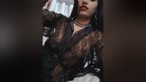 Media: A video of a woman with long black hair, wearing a sheer black lace robe, standing against a blurred background. She has a light to medium skin tone, a slender figure, and is adorned with a pearl necklace.