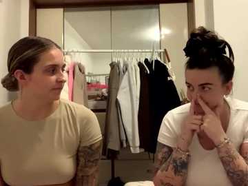 Media: Video of two women with light skin, one in a beige top, the other in a white top, standing in a wardrobe with hanging clothes, whispering secrets.