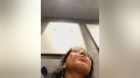 Media: A video of a young woman with medium skin tone, wearing glasses and black hair, looking upward in a dimly lit, modern interior with rectangular lights and grey walls.