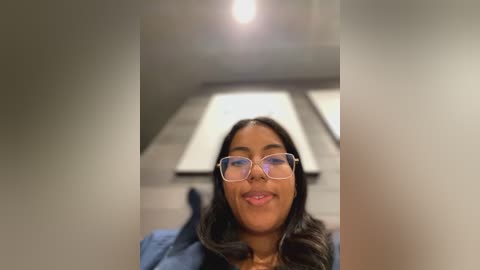 Media: Video of a smiling, light-skinned woman with glasses, medium-length black hair, wearing a blue top, lying on a gray carpeted floor with a ceiling light overhead.