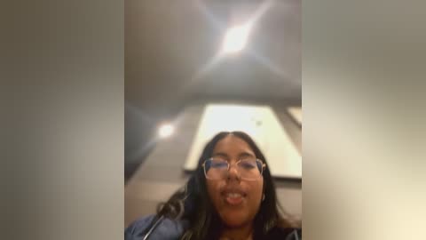 Media: A video of a young woman with medium-brown skin, long dark hair, and wearing glasses, smiling, standing indoors with a modern, minimalist ceiling.