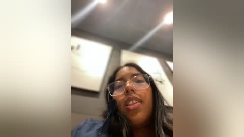 Media: A video of a Black woman with medium-brown skin, wearing glasses and a blue jacket, leaning forward with a slight smile, taken in a dimly lit room with recessed ceiling lights and a blurred background.