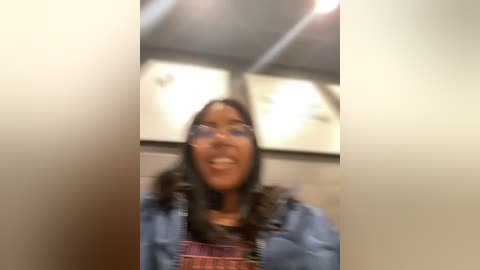 Media: Video of a smiling young woman with medium brown skin, long dark hair, wearing glasses and a denim jacket over a red and black striped shirt. Background features a beige tiled wall and fluorescent lights.