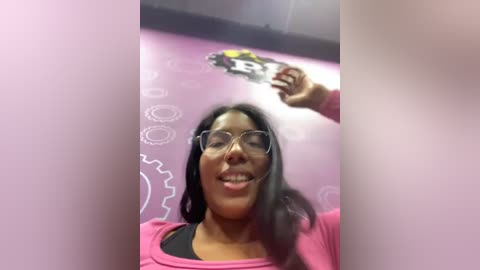 Media: A video of a smiling woman with long black hair and glasses, wearing a pink top, holding a football. The background features a pink wall with white gear and gear icon designs.