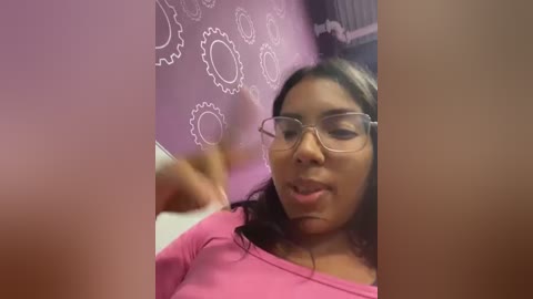 Media: A video of a young woman with medium brown skin, wearing glasses and a pink shirt, holding a pink phone in a room with a purple wall adorned with white gears.