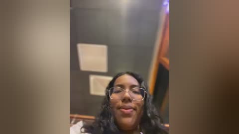 Media: Video of a young Black woman with curly hair and glasses, smiling, taken in a dimly lit room with abstract art on the walls.