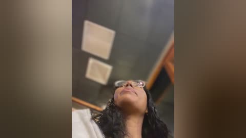 Media: A video shows a young woman with long black hair, wearing glasses, looking upwards. She is in a dimly lit room with a chalkboard and rectangular windows in the background.