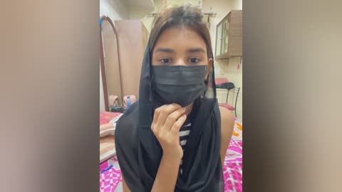 Media: Video of a young woman with medium brown skin, wearing a black mask, a black headscarf, and a black shirt. She is in a small, cluttered bedroom with pink and white bedding, a mirror, and a wooden cabinet.