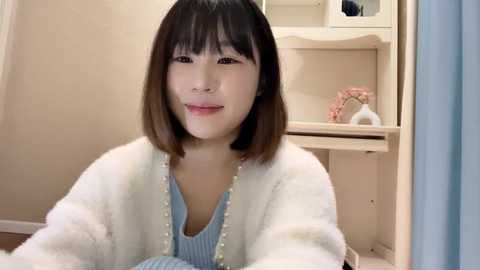 Media: Video of an East Asian woman with straight, shoulder-length brown hair, wearing a white fur cardigan over a light blue sweater. She sits in a cozy, light-colored room with a white dresser and floral decor in the background.