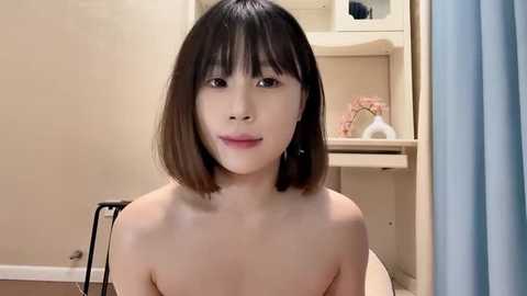 Media: Video of a topless East Asian woman with shoulder-length black hair, fair skin, and a neutral expression. Background features beige walls, a chair, and a shelf with decorative items.