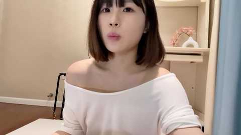 Media: A video of a young East Asian woman with straight, shoulder-length brown hair and fair skin, wearing a white off-shoulder top. She sits in a room with beige walls and wooden furniture.