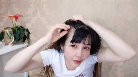 Media: Video of a young Asian woman with straight black hair, wearing a white T-shirt, combing her hair in front of a beige wallpaper with a floral pattern.