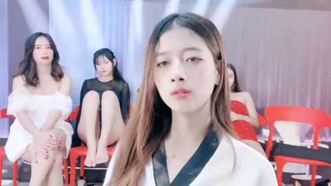 Media: Video of four East Asian women, including a close-up of a woman with long brown hair in a white robe, seated on red chairs in a modern, brightly lit room with white curtains.