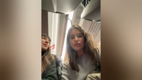 Media: Video of two women, one with long brown hair and a gray sweater, the other with long blonde hair and a green jacket, seated in a train compartment with beige walls and a window.