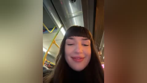 Media: A video of a smiling young woman with long, straight brown hair and bangs, wearing a black top, taken in a bus with metallic ceiling and yellow handrails.