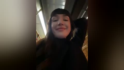 Media: A video of a young Asian woman with straight black hair, smiling, wearing a black turtleneck sweater, standing in a dimly lit subway car.