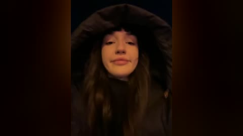 Media: A video of a young woman with long brown hair, wearing a dark hooded jacket, set against a dark background with warm orange lighting.