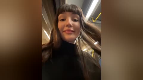 Media: Video of a young woman with straight brown hair, blunt bangs, and fair skin, wearing a black turtleneck sweater, smiling. She's in a dimly lit subway car with yellow lines on the walls, looking into the camera.