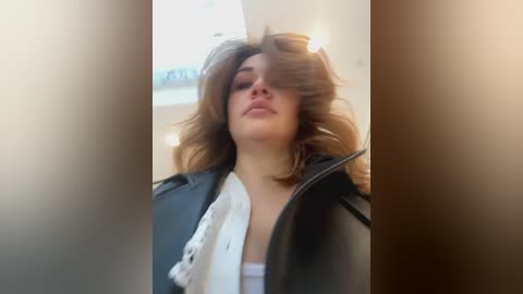 Media: A video of a young woman with wavy, shoulder-length brown hair, wearing a black leather jacket and white top, taken indoors with soft lighting, creating a blurred, dreamy effect.