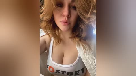 Media: A video of a young, fair-skinned woman with wavy, shoulder-length auburn hair, wearing a white Hooters tank top revealing cleavage, taken indoors with soft lighting.