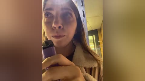 Media: Video of a young woman with light brown skin and long brown hair, holding a purple hair straightener, wearing a beige fur-trimmed coat indoors.