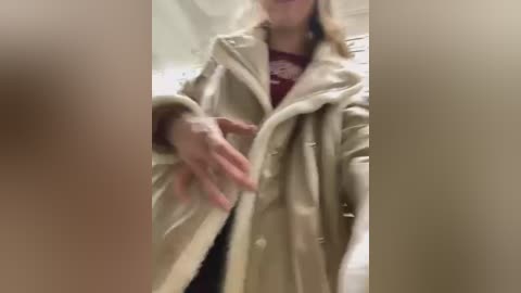 Media: A blurry video shows a person with light skin and blonde hair wearing a beige leather jacket over a maroon top. Their hands are adjusting the jacket\u2019s buttons. The background is indistinct, likely indoor.