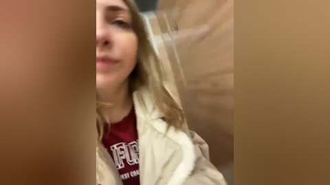 Media: A blurry video of a young woman with long blonde hair and fair skin, wearing a red t-shirt and beige jacket, taken indoors with out-of-focus objects in the background.