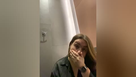 Media: Video of a woman with long brown hair, wearing a black jacket and a watch, covering her face with her hands, standing in a bathroom with beige and white tiled walls and a metal hook.