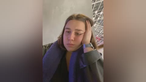 Media: A video of a young woman with shoulder-length brown hair, wearing a gray coat and a brown top, looking tired and holding her head, in a dimly lit room with metal structures and a concrete wall.