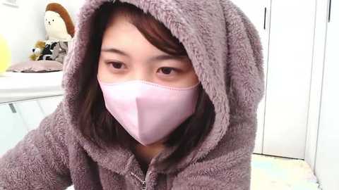 Media: Video of an Asian woman with straight brown hair wearing a light pink face mask and a soft, grey hooded robe, in a modern, white-walled bathroom with a white sink and a plush, stuffed toy.