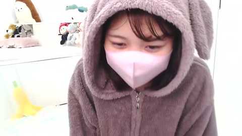 Media: A video of an Asian child in a grey bunny-themed hooded robe, wearing a pink surgical mask, standing indoors with a white shelf and plush toys in the background.