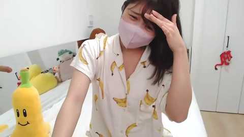 Media: Video of an Asian woman in a white shirt with yellow bananas, wearing a mask, scratching her head, in a bright, white room with plush toys and a toy banana.