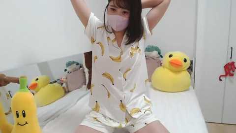 Media: Video of a young East Asian woman with fair skin and straight black hair, wearing a white shirt with yellow bananas and shorts, posing on a bed with yellow duck toys and a banana-themed pillow.