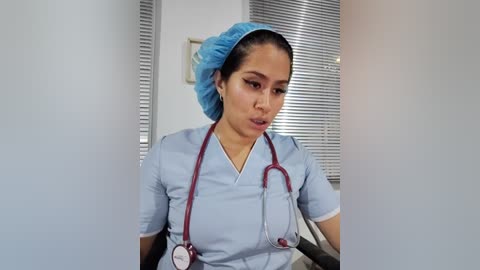 Media: Video of a Latina woman with medium skin tone, medium build, and medium-sized breasts, dressed in light blue scrubs and blue cap, holding a stethoscope, in a hospital room with closed blinds.