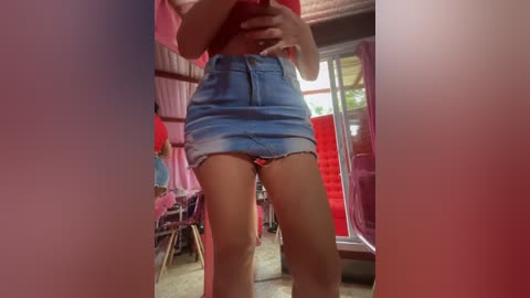 Media: Video of a woman in a red top and denim shorts standing in a dimly lit room with pink walls and red furniture, taking a selfie with a red phone.