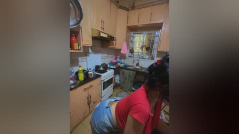 Media: Video of a cluttered, outdated kitchen with light wood cabinets, a stove, and a sink. A person in a red shirt and jeans is crouched on the floor, possibly cleaning.