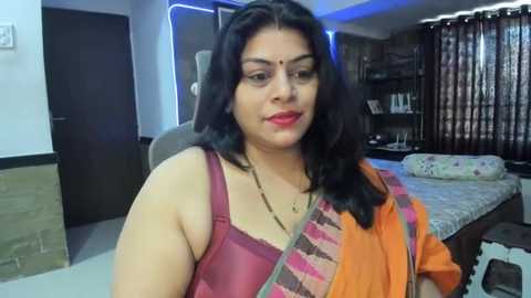 Media: Video of a South Asian woman with medium skin tone, wearing an orange saree with a maroon blouse, sitting in a modern bedroom with blue lighting and a bed with floral sheets.