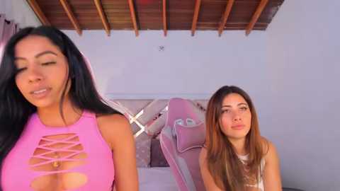 Media: Video of two women in a room: One with long black hair in a pink crop top, the other with long brown hair in a white top. Wooden ceiling, gaming chair in background.