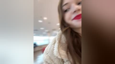 Media: Video of a smiling woman with long brown hair, wearing a beige fuzzy sweater, standing in a modern, brightly lit indoor space with white walls and recessed lighting.